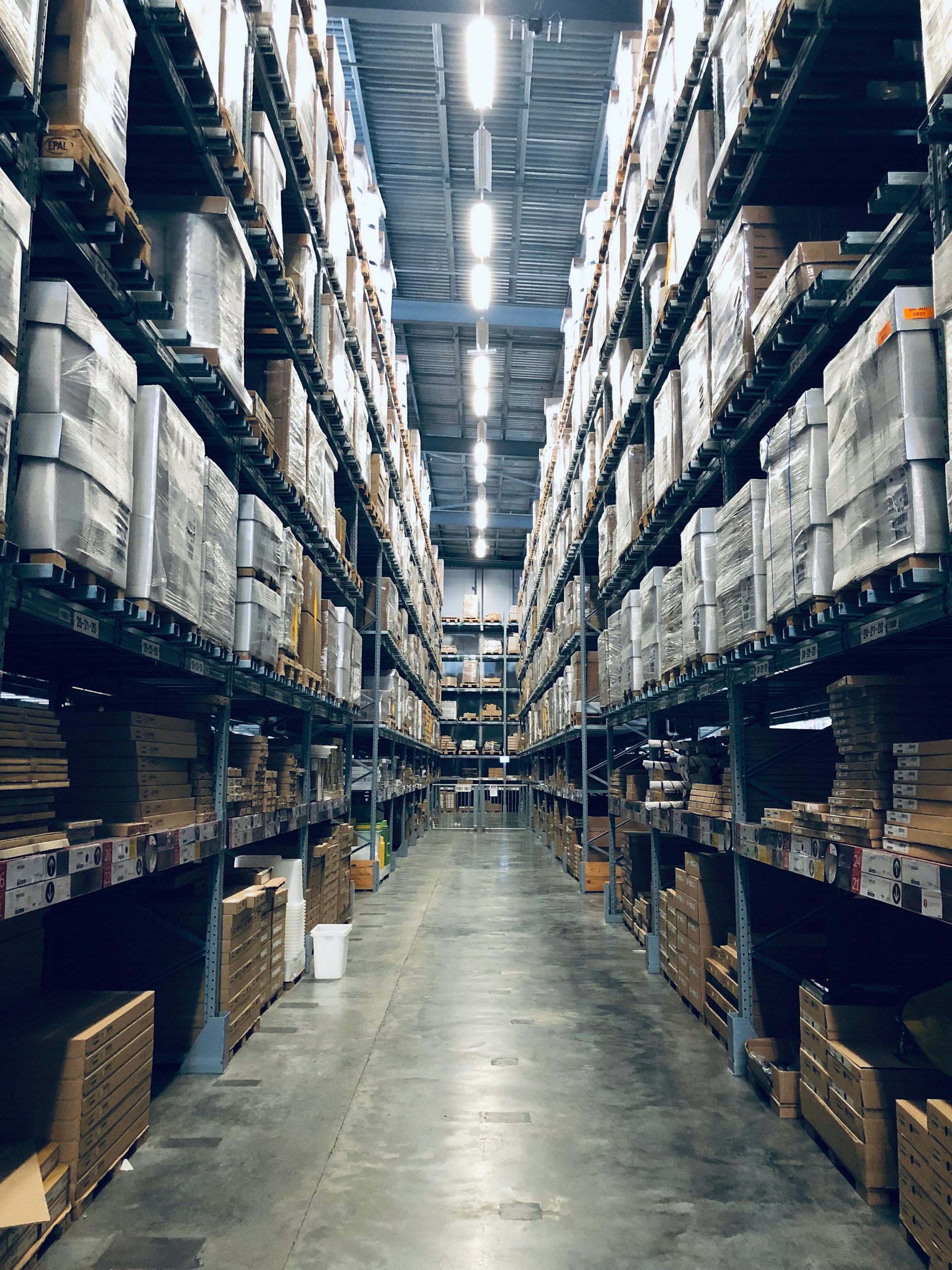 A long aisle inside a high-bay warehouse lined with tall steel racking under bright overhead lighting.